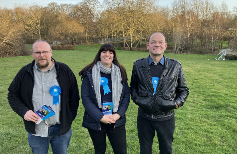 David, Victoria and Matthew stood facing the camera wearing blue Conservative rosettes. In the background is a meadow and woodland.
